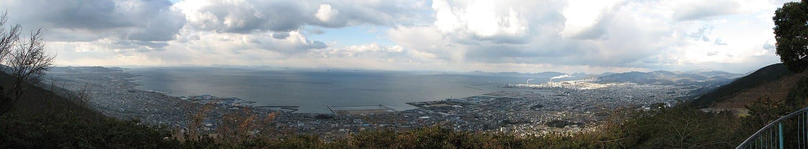 A wide panorama of a coastal town and a calm sea under clouds, seen from a hillside